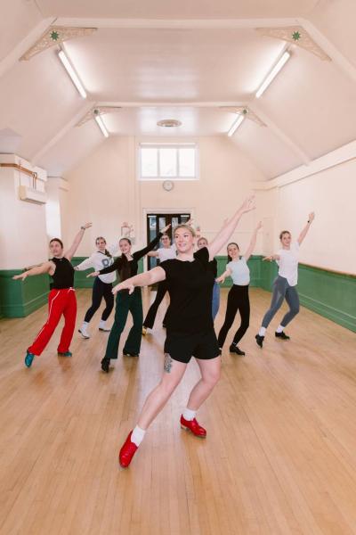 A dance teacher dressed in black tshirt and shorts wearing red tap shoes poses at the front of a tap class. Teacher and dancers are posing with their left leg bent, right let outstretched, and arms lifted,