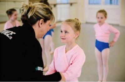 Ballet teacher in a black jacket, facing a group of young dancers in ballet attire. The teacher is holding one student's hands.